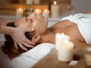 una donna su un lettino con asciugamano e candele intorno viene massaggiata sulle tempie